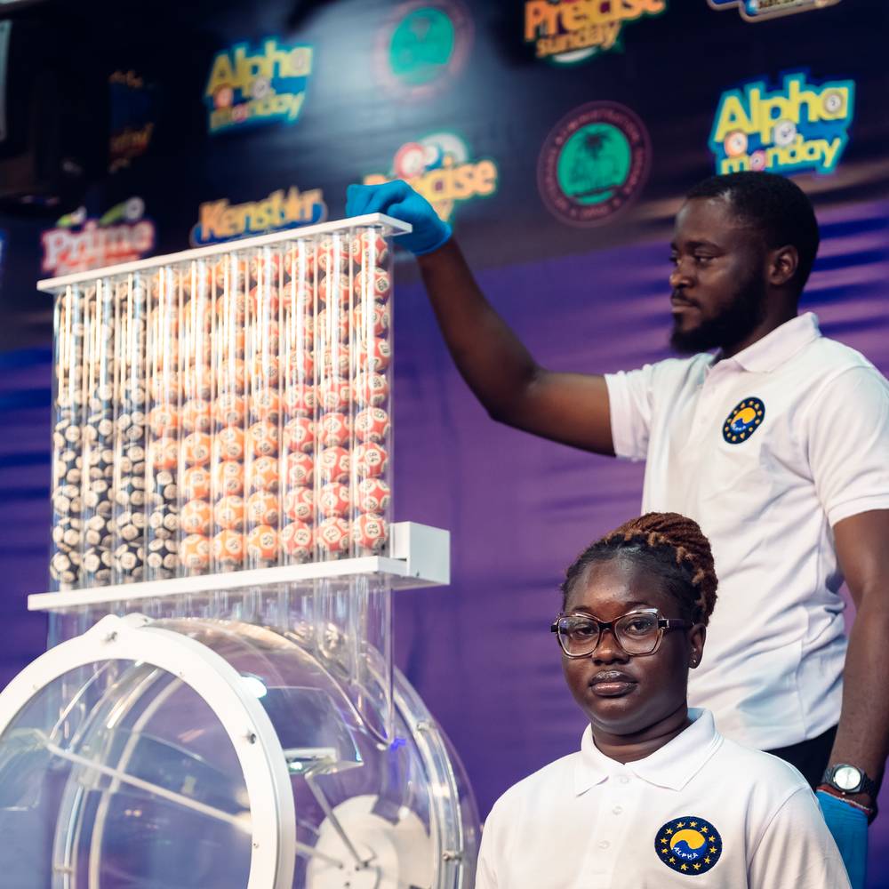 Two Alpha Lotto draw officers in white uniform at a draw machine, one loading the rack of numbered balls