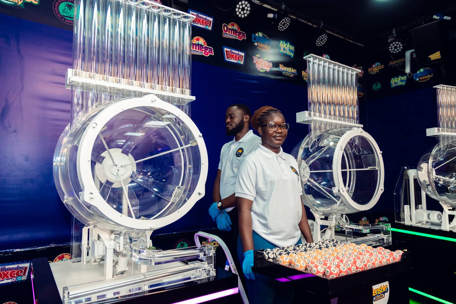 Two Alpha Lotto draw officers in white uniform standing at a draw machine with a tray of numbered balls