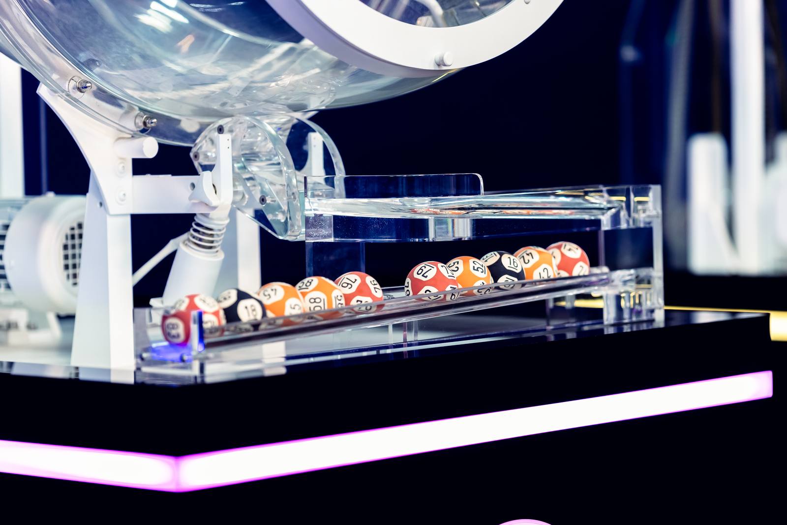 Drawn balls resting in the clear tray beneath a draw machine, lit by the plinth's neon strip